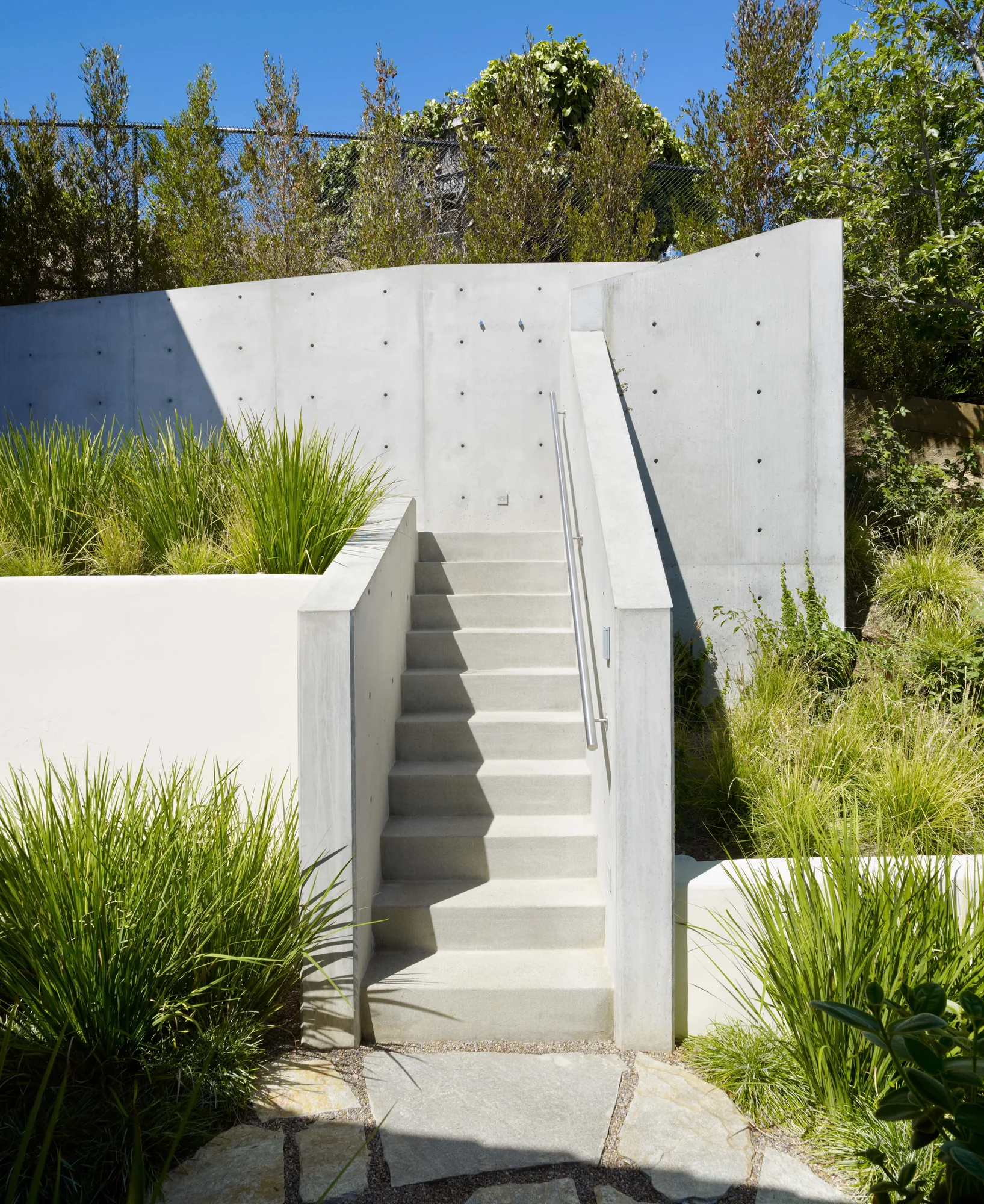 Concrete staircase ascending through modern landscape with ornamental grasses and retaining walls under blue sky