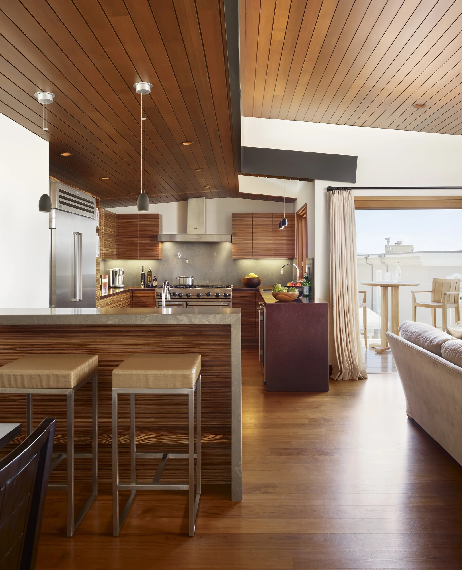 Modern kitchen with wood slatted ceiling, stone counter island with stools, and view to outdoor patio