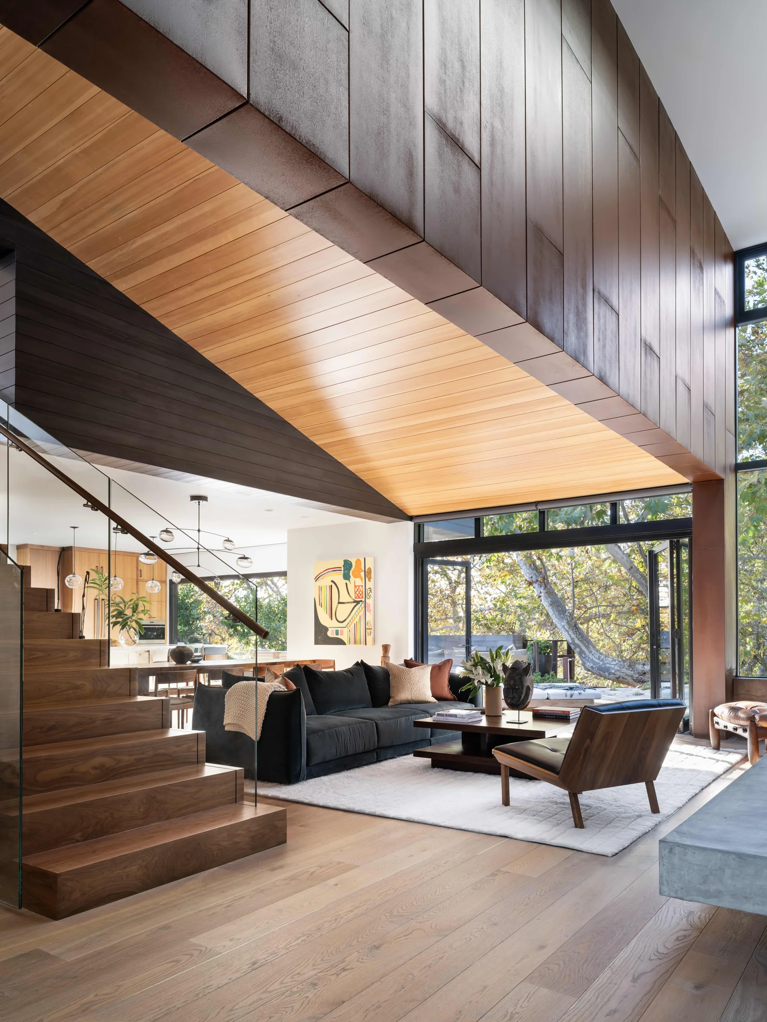 Modern living room with vaulted wood ceiling, floor-to-ceiling windows, and open staircase overlooking natural landscape.