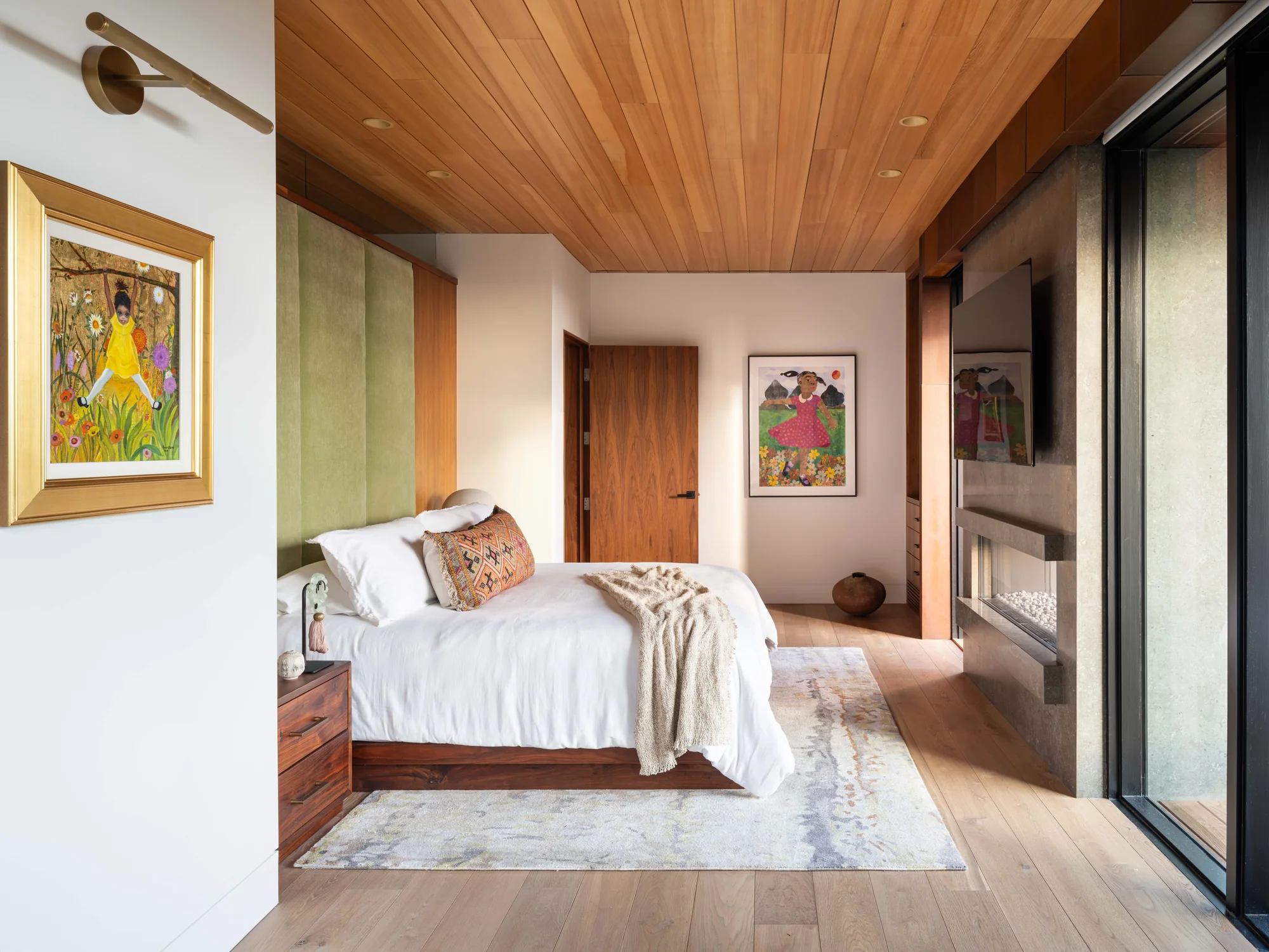 Modern bedroom with wood ceiling, white bedding, gold-framed artwork, and glass doors opening to outdoor space.