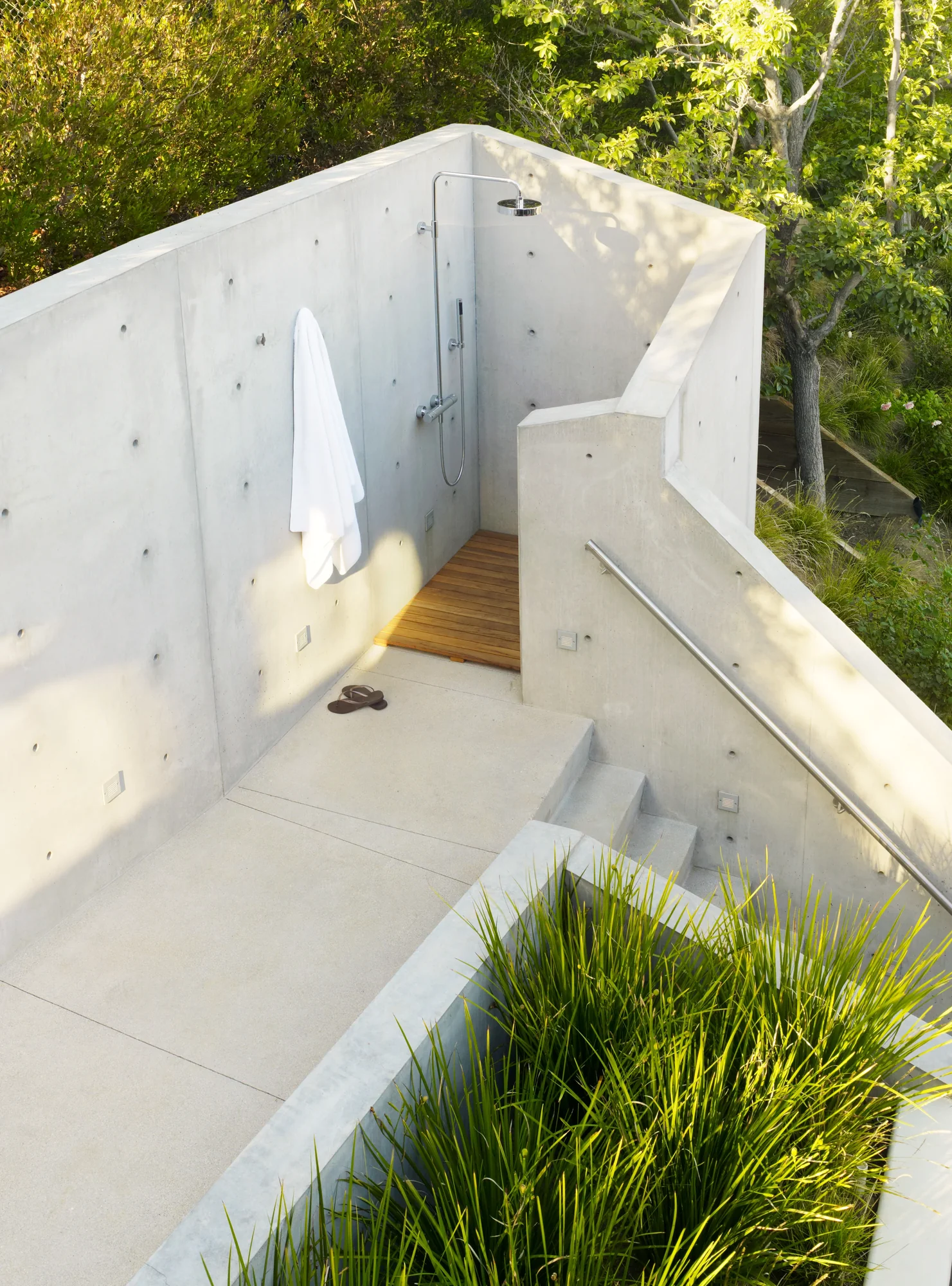Concrete outdoor shower surrounded by lush greenery, featuring wood mat, modern fixtures, and integrated seating.