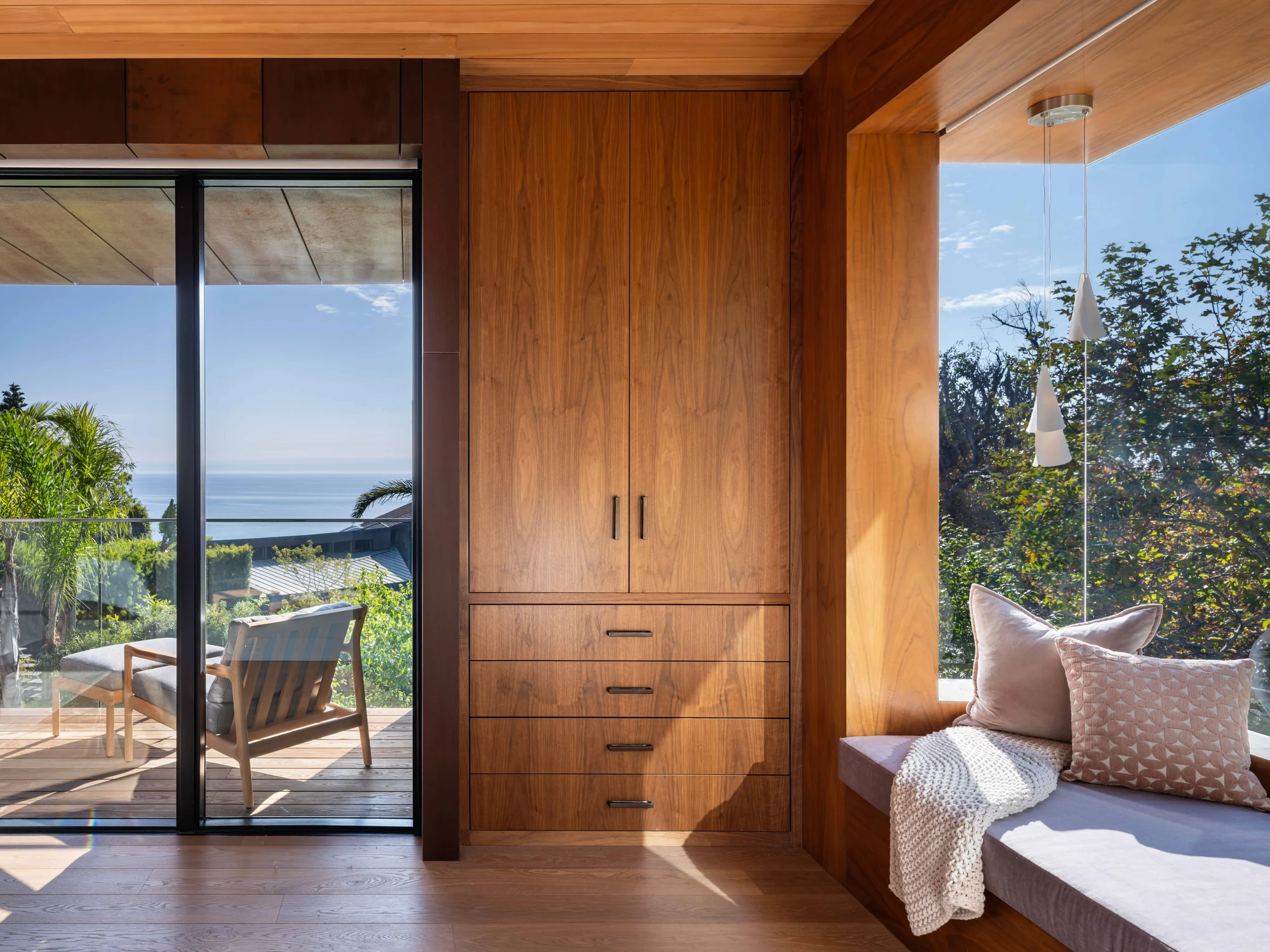 Modern bedroom with built-in walnut wardrobe, window seat with pink pillows, and glass doors opening to ocean-view deck