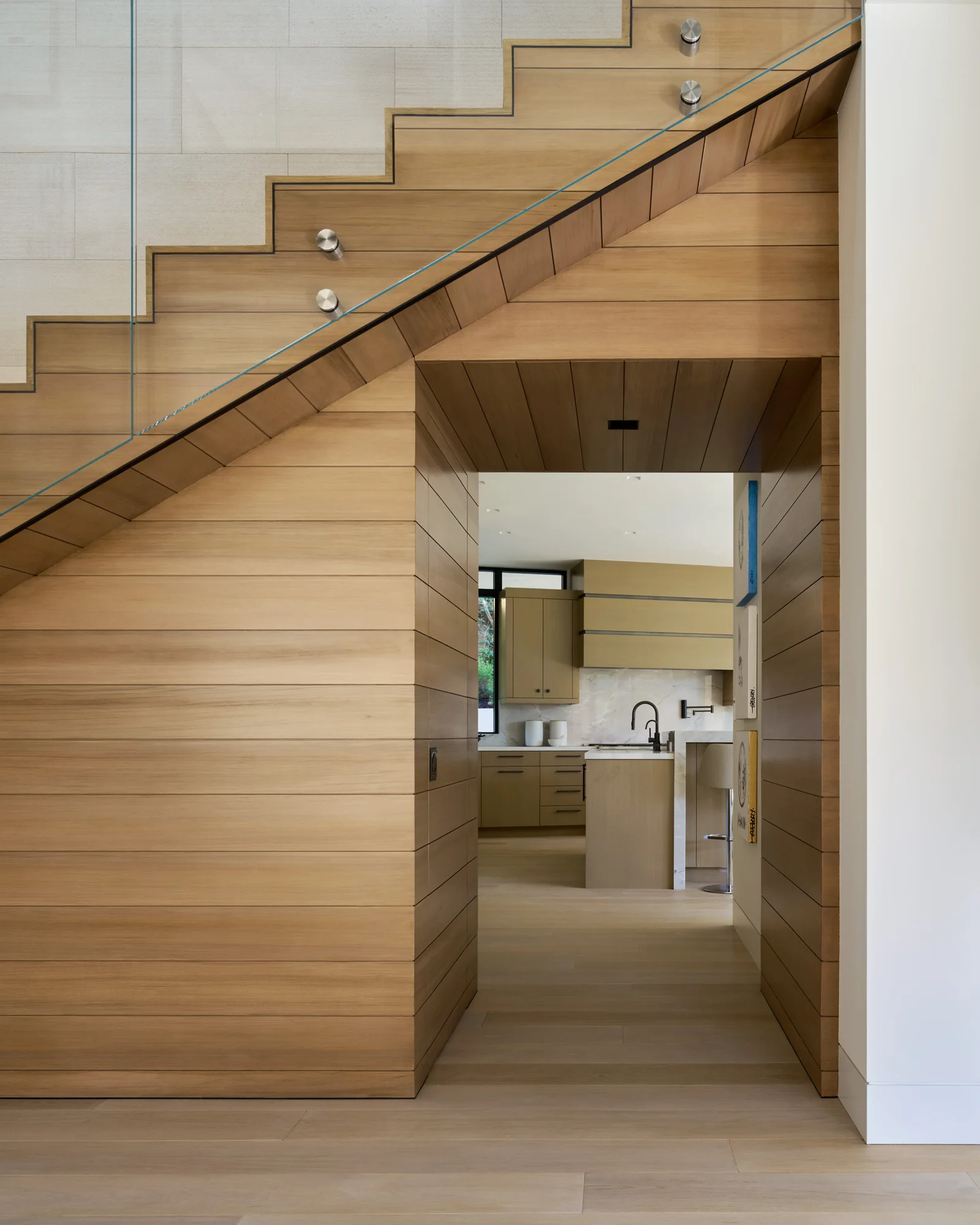 Cedar-clad staircase with glass railing flows over a wood-paneled passage leading to a modern kitchen beyond.
