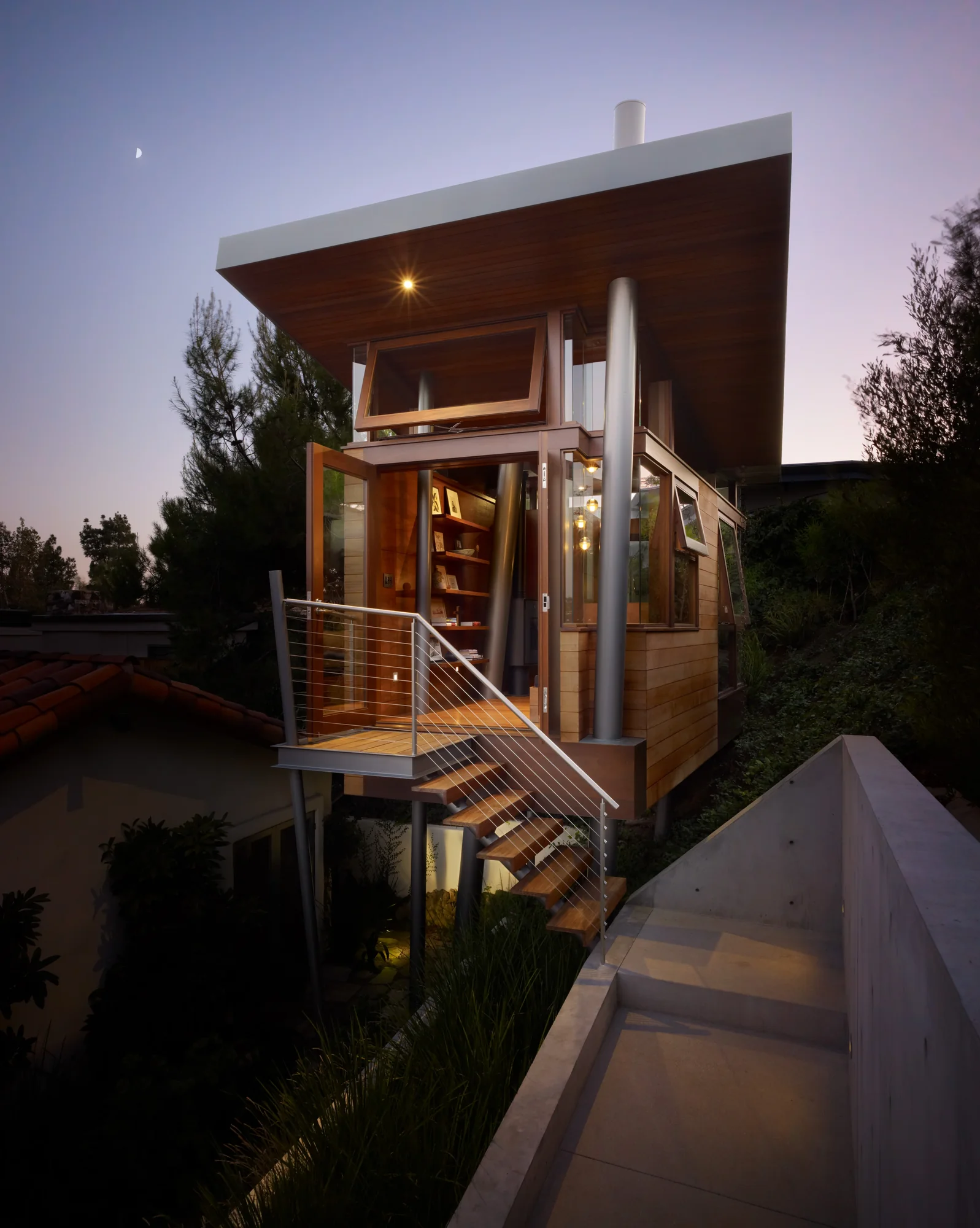 Modern elevated wood and glass studio with steel supports at twilight, featuring cable railing stairs and natural landscaping.