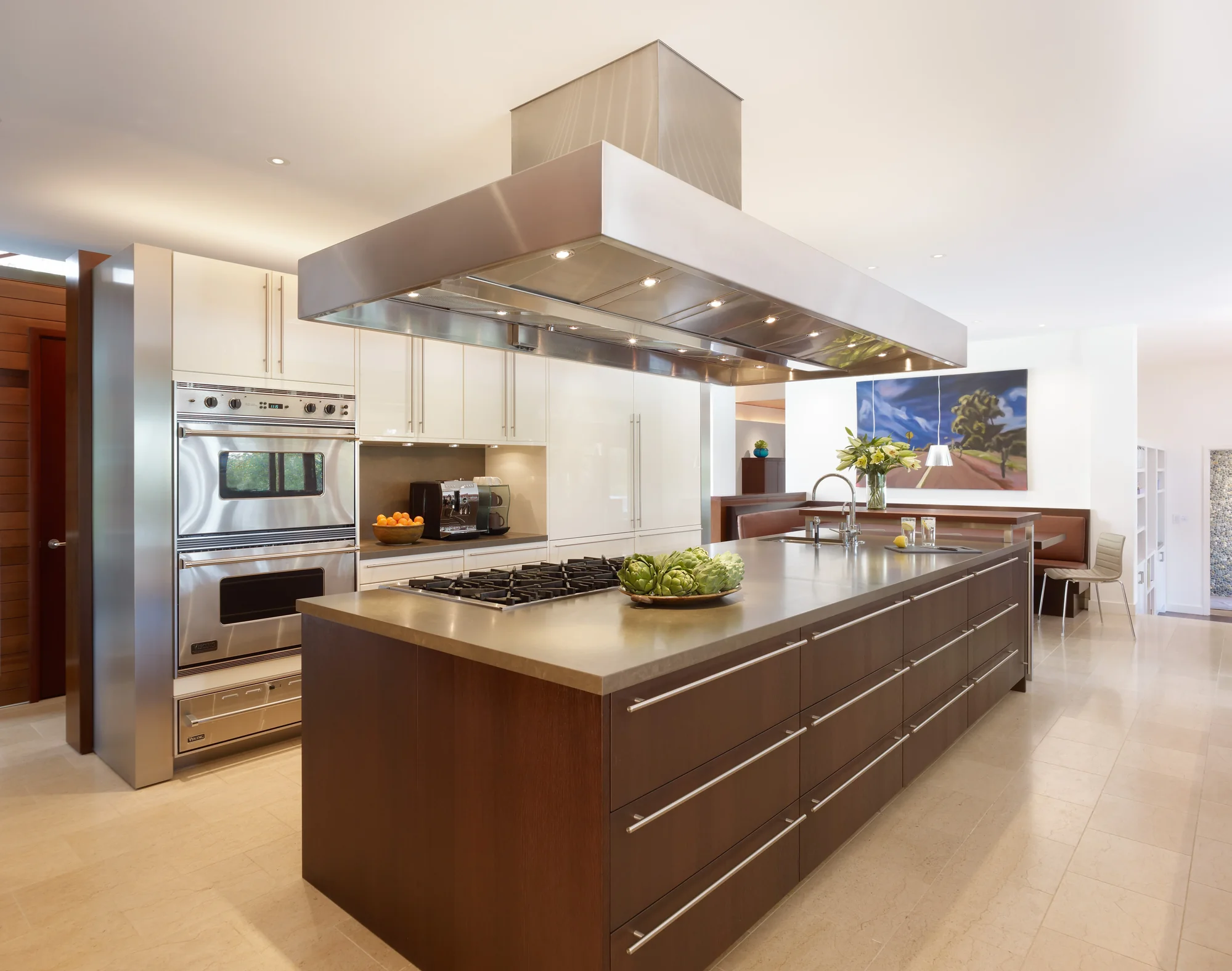 Modern kitchen with large brown wood island, gas cooktop, stainless steel hood, white upper cabinets, and built-in ovens