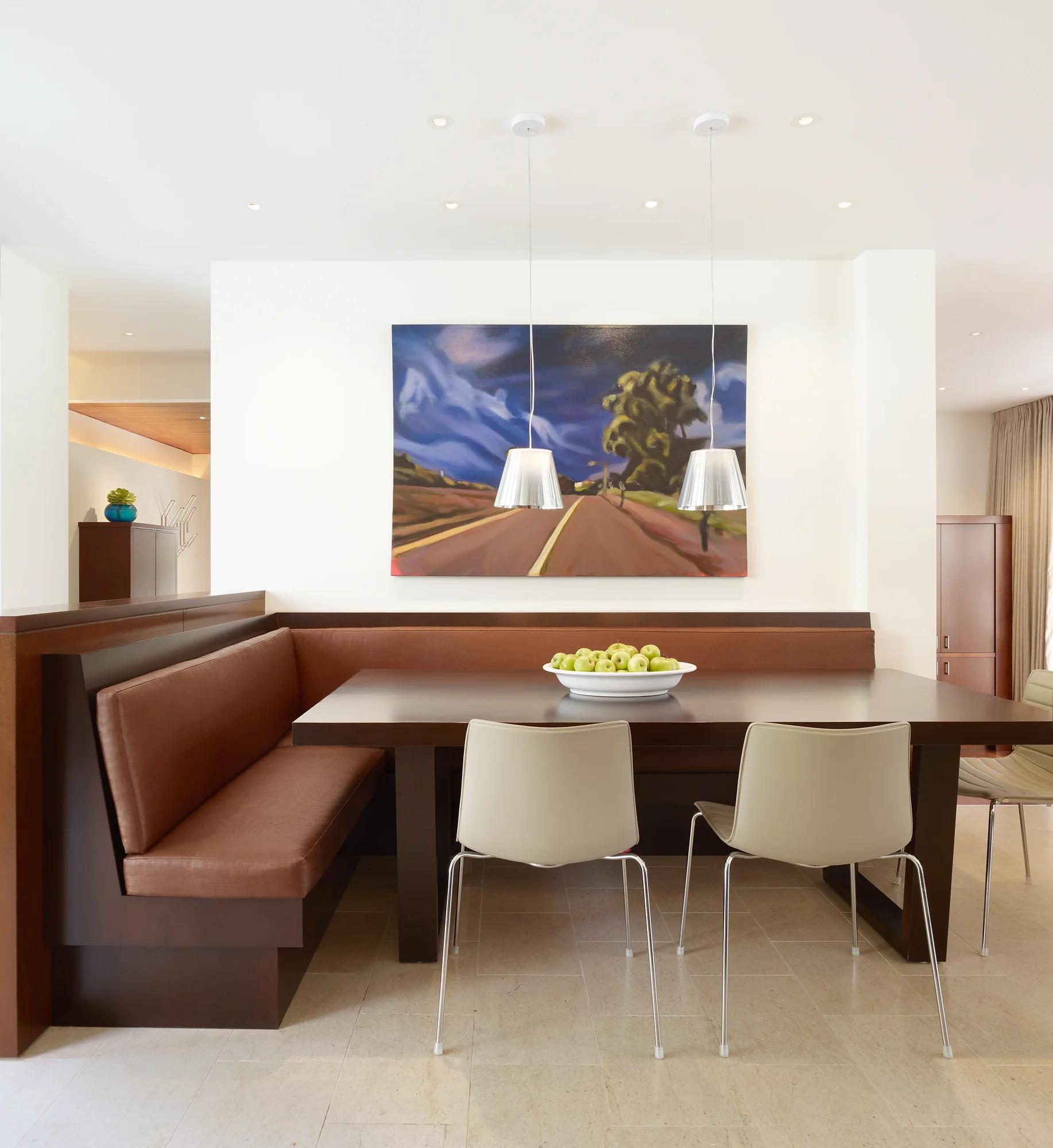 Modern dining nook with mahogany banquette, stone tile floor, pendant lights, and landscape painting on white wall.