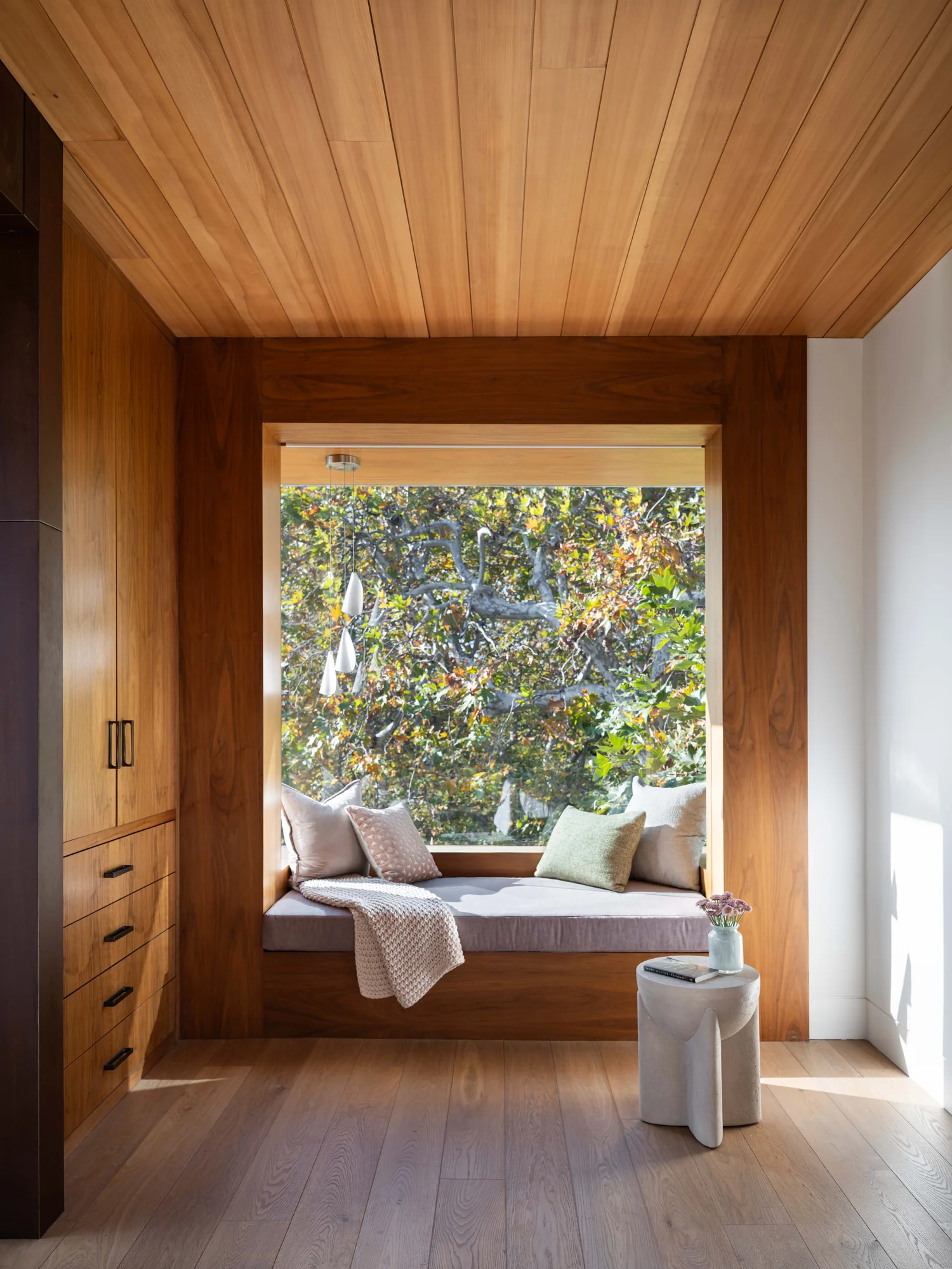 Cozy window seat nook with wood paneling, cushions, and view of trees, featuring built-in storage and natural light.