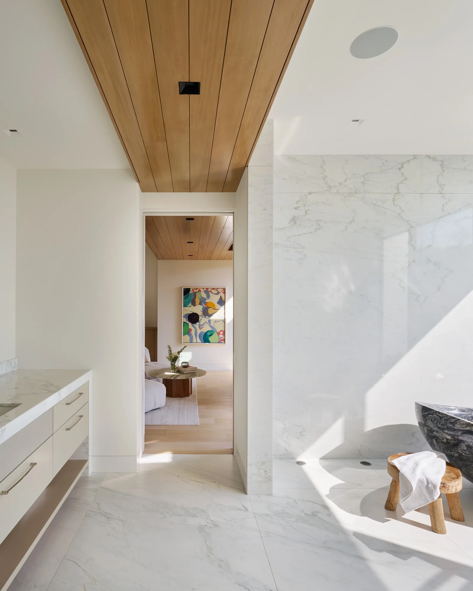 Luxurious master bathroom with marble floors, wood-slat ceiling, freestanding stone tub, and open view into serene bedroom beyond.