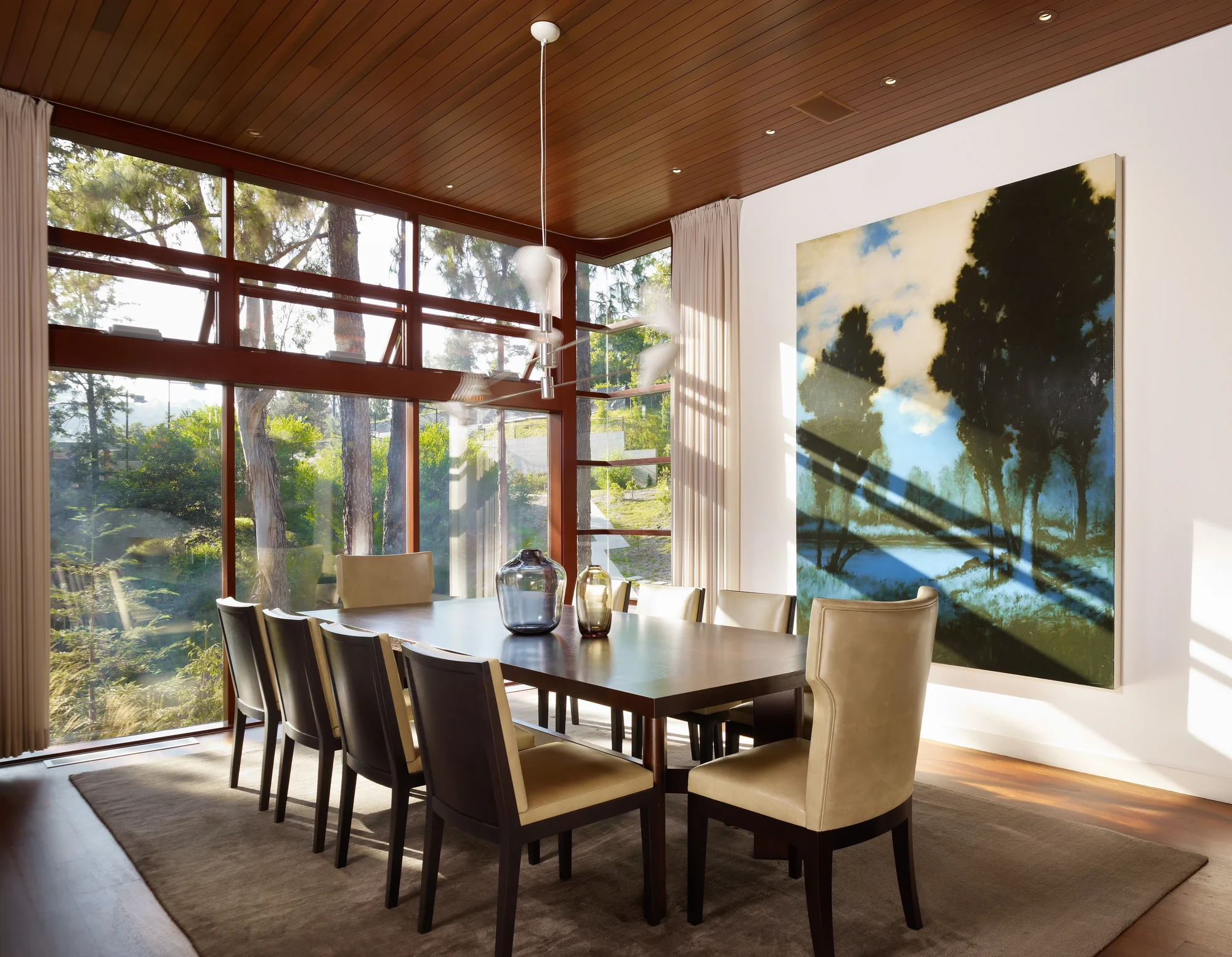 Dining room with mahogany ceiling, floor-to-ceiling windows overlooking lush trees, dark wood table, and large landscape painting.