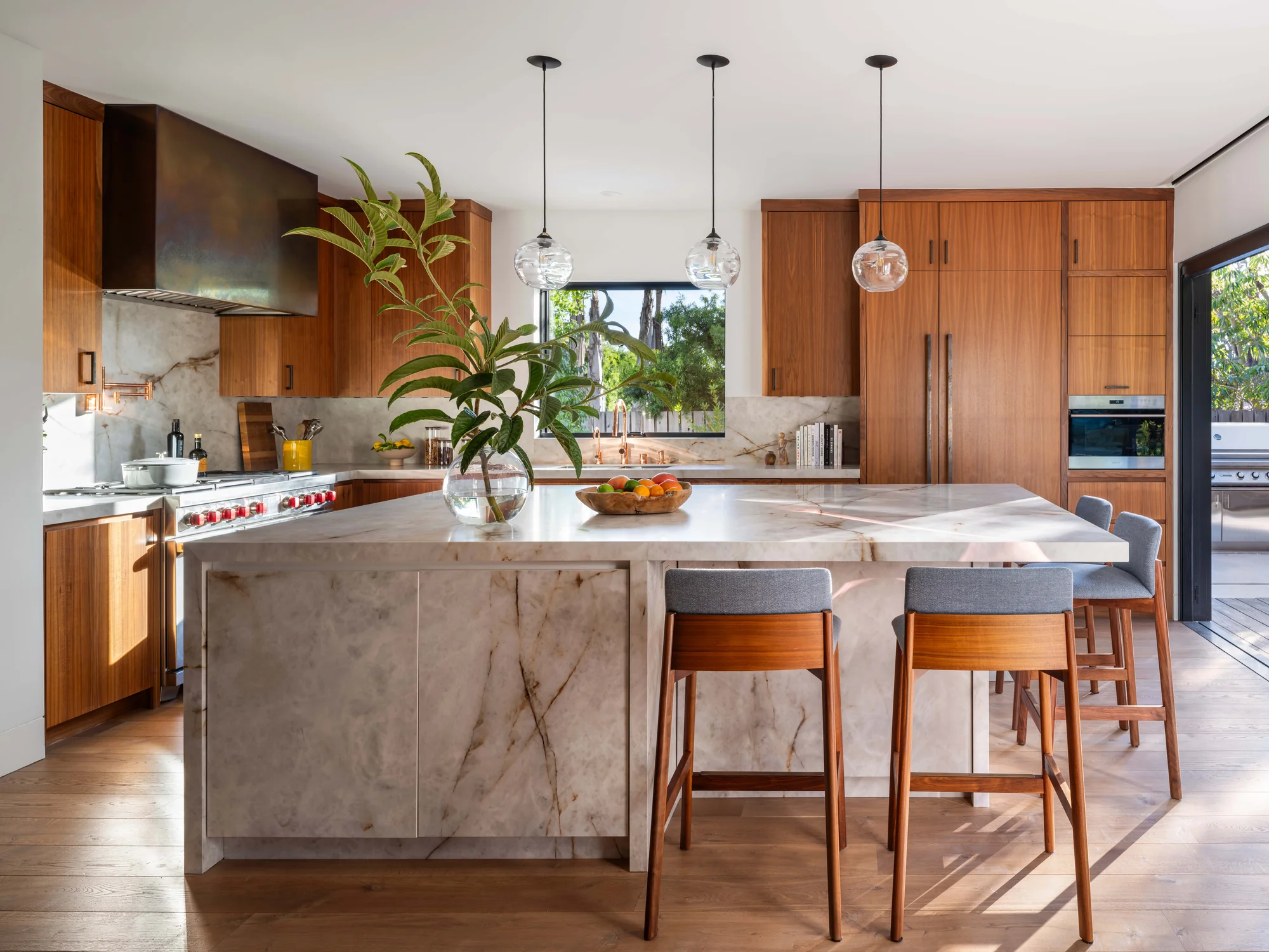 Modern kitchen with marble island, warm wood cabinetry, pendant lights, and garden views through black-framed windows.