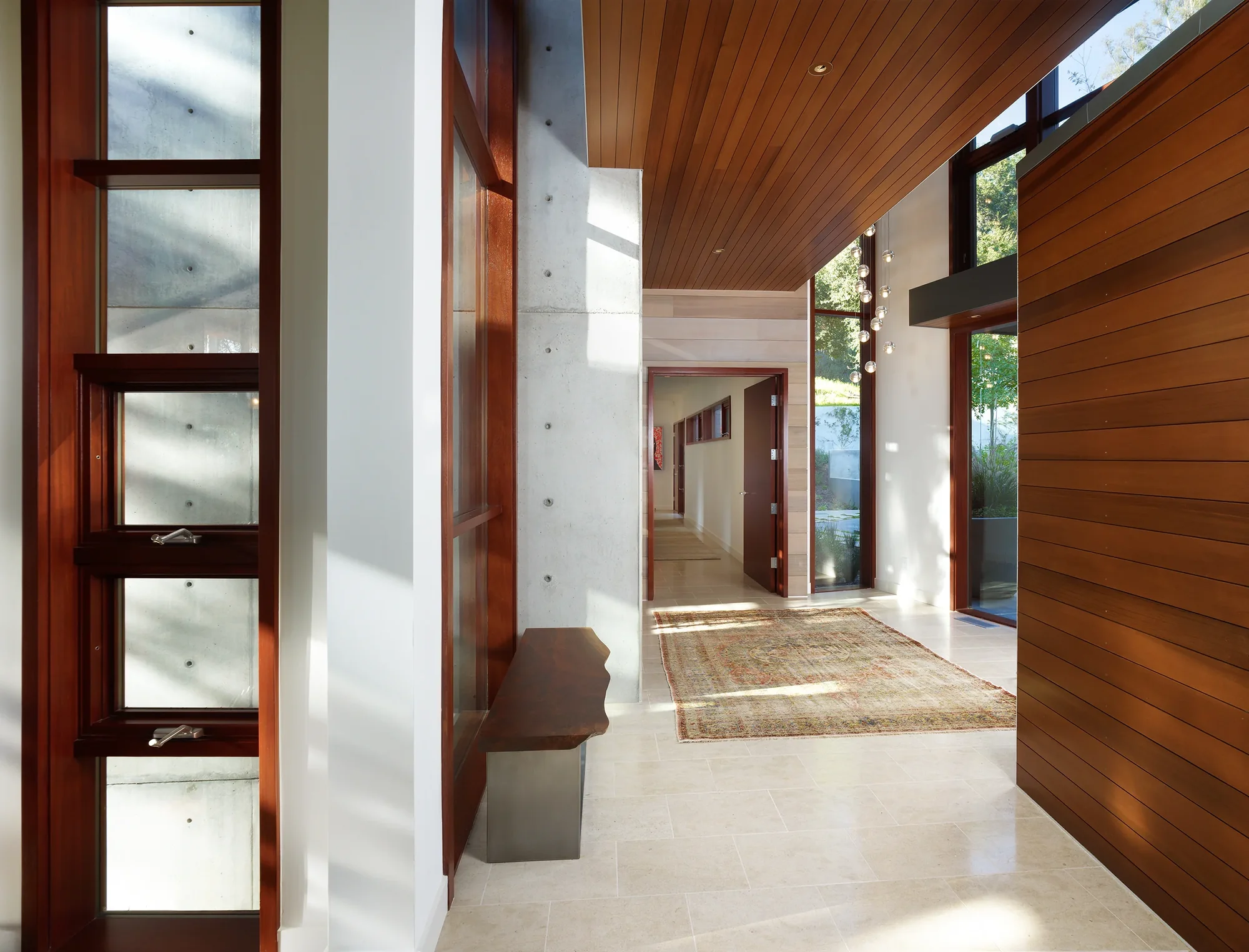 Modern entryway with wood pivot door, concrete walls, cedar ceiling, and stone flooring connecting indoor-outdoor spaces