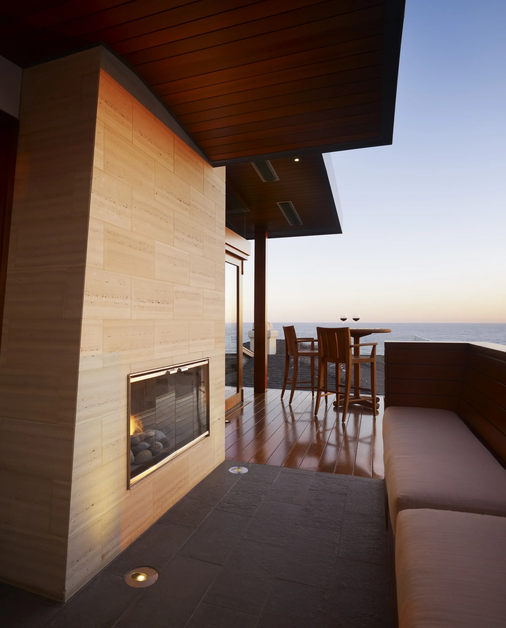 Modern outdoor deck with travertine fireplace, wooden dining area, and ocean views at dusk