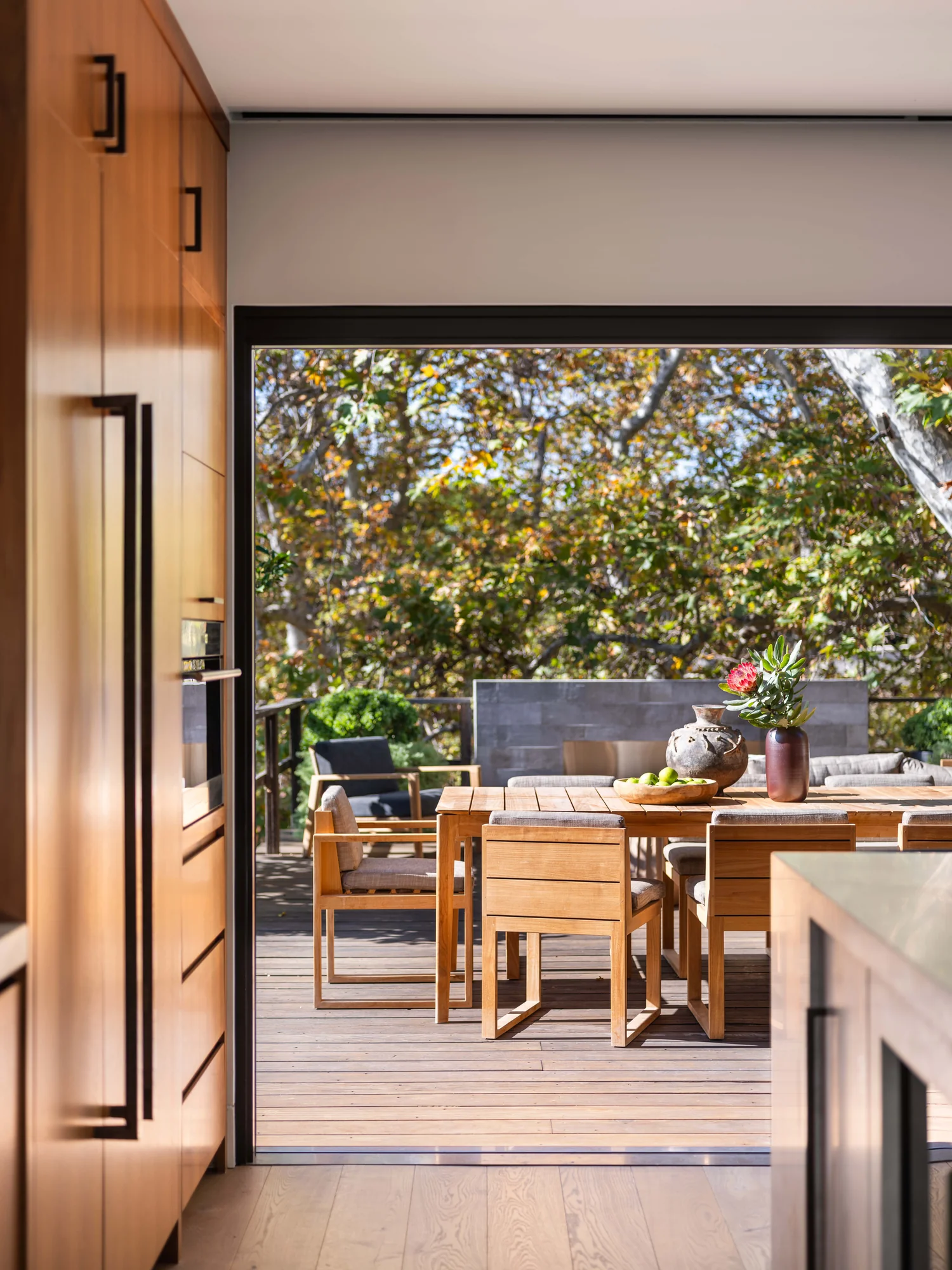 View from interior through open sliding doors to wooden deck with dining table overlooking tree canopy