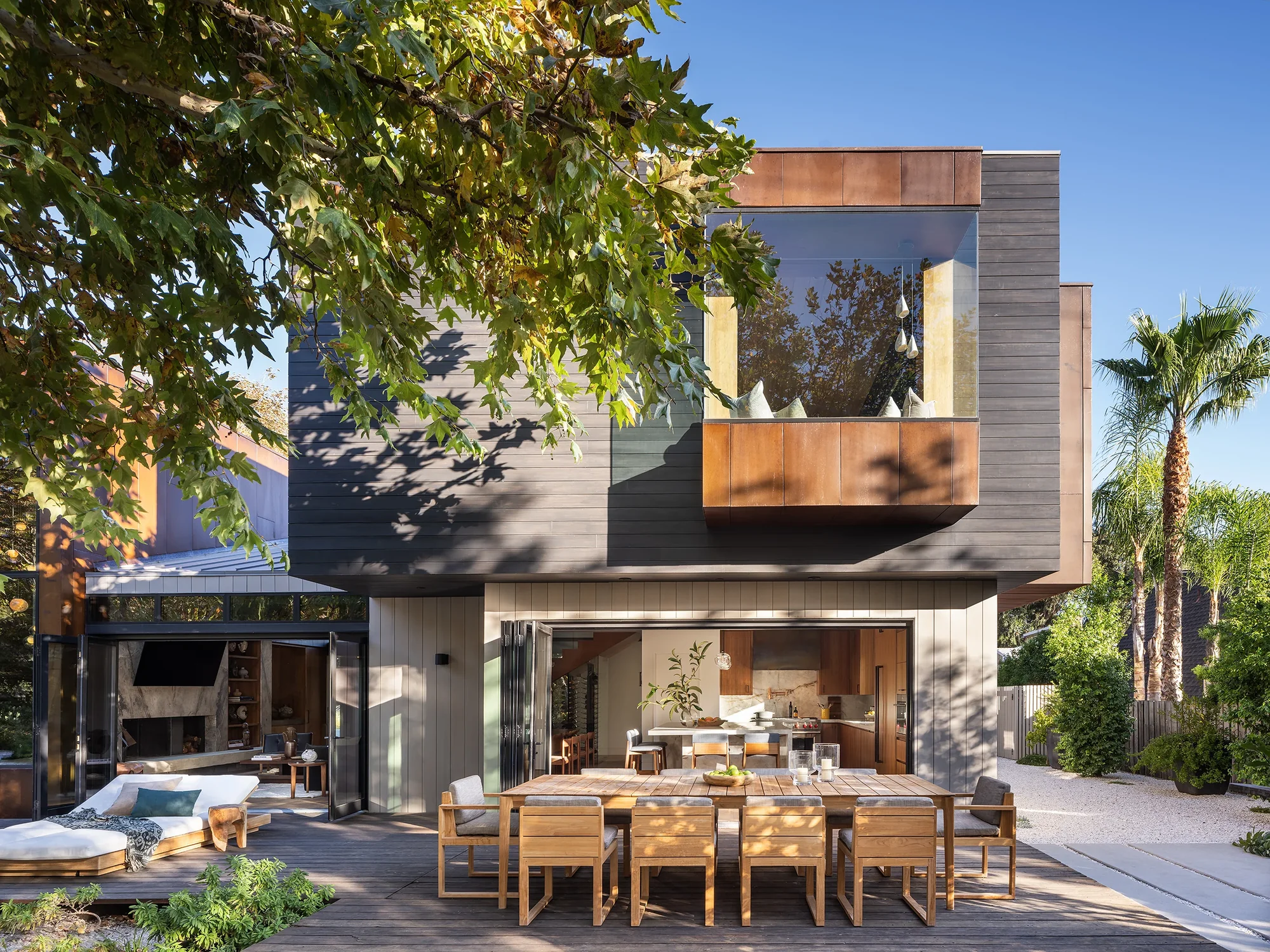 Modern two-story home with copper accents featuring an outdoor dining area connecting to an open kitchen under tree-shaded patio.