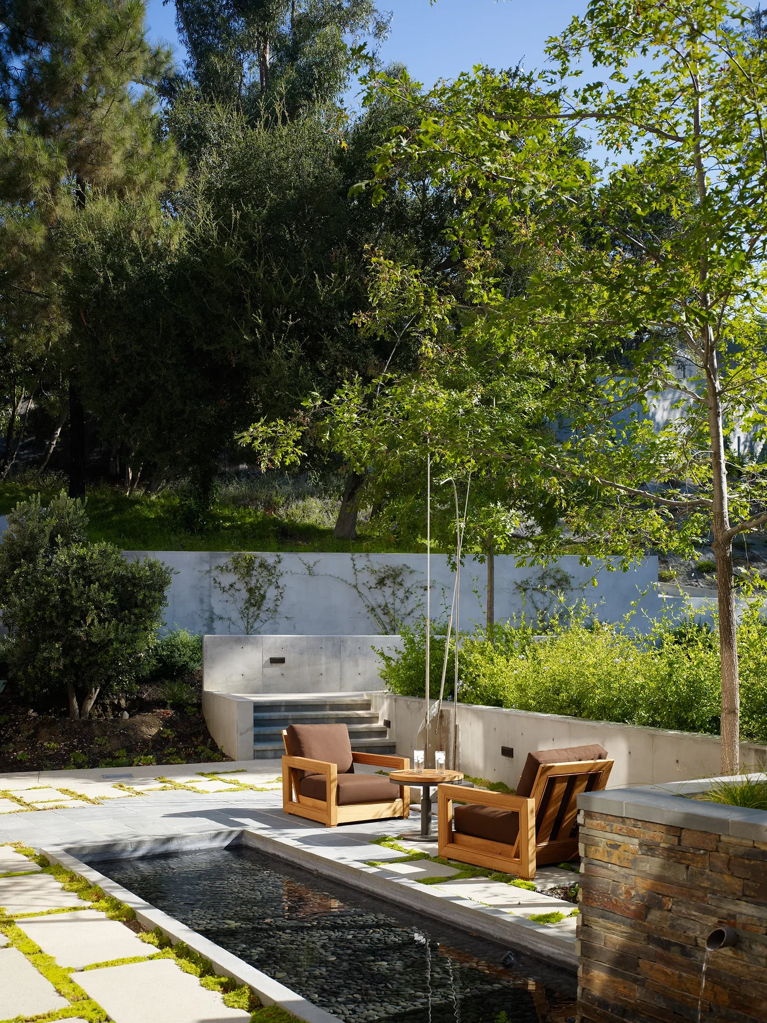 Terraced outdoor patio with teak lounge chairs beside a reflecting pool, concrete retaining walls, and lush hillside landscaping.
