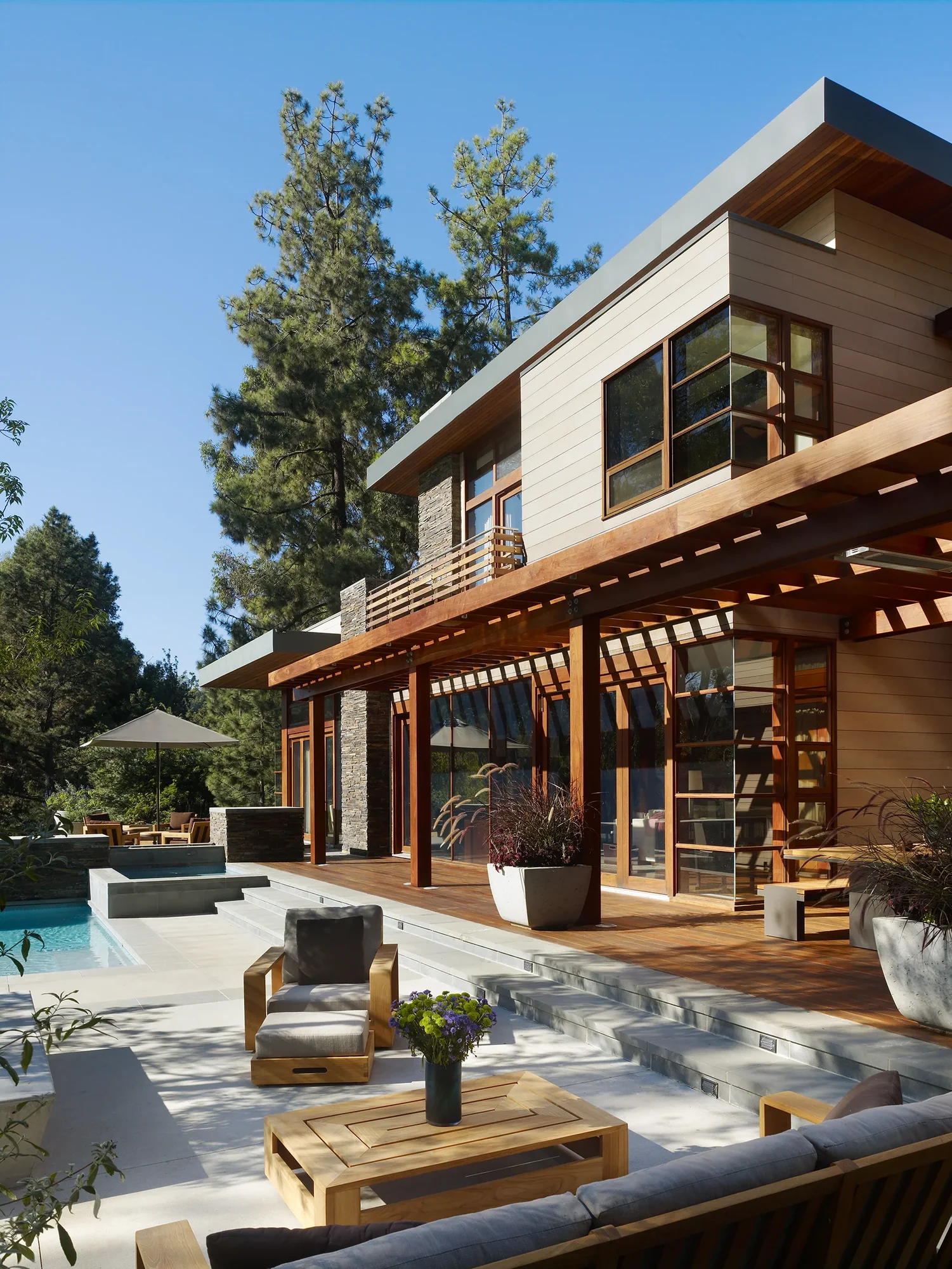 Exterior patio of a modern custom home with cedar pergola, stone accents, pool, and teak outdoor furniture amid tall pines.