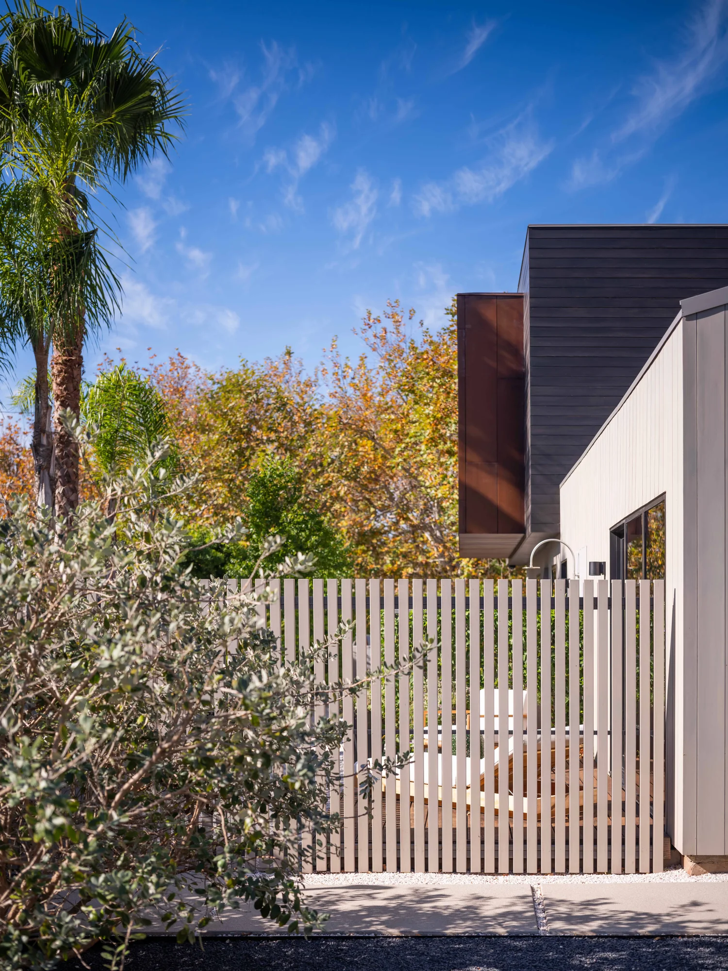 Modern home entry with vertical wood slat fence, dark horizontal siding, and lush landscaping under blue sky.