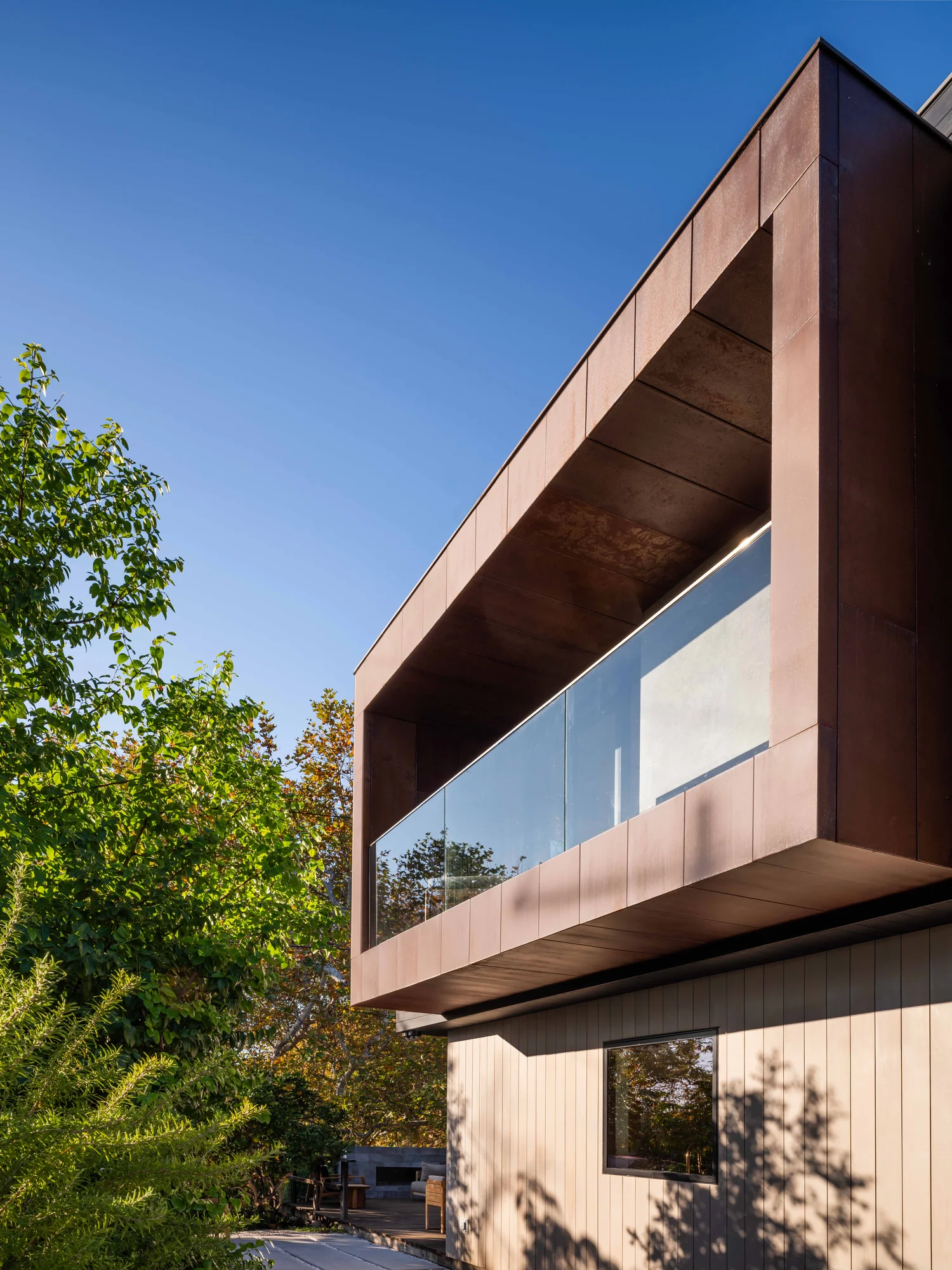 Modern two-story home with cantilevered upper level clad in copper panels and cedar siding below, framed by trees.