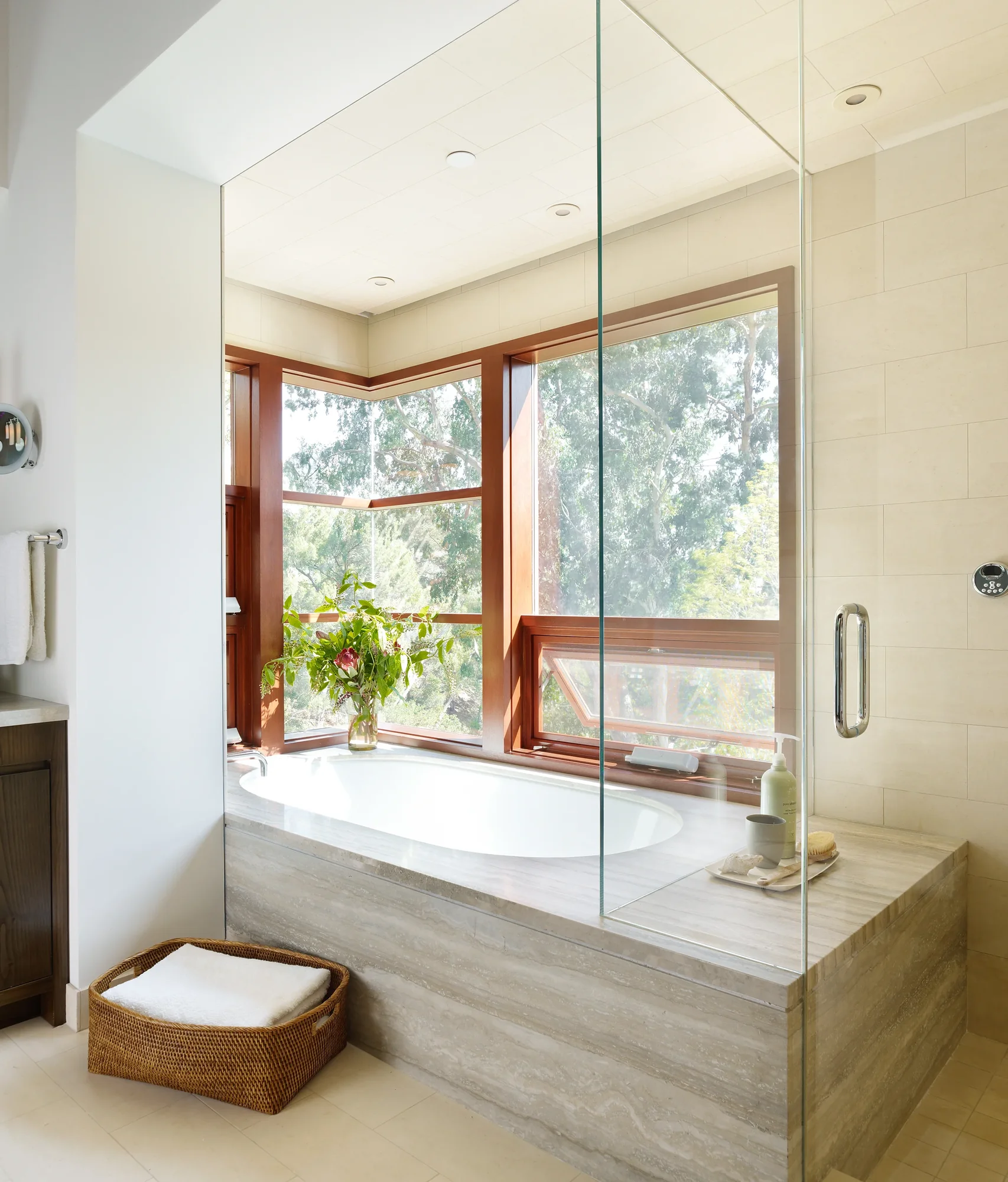 Marble-clad soaking tub beneath corner mahogany-framed windows overlooking lush tree canopy, with adjacent glass shower enclosure.
