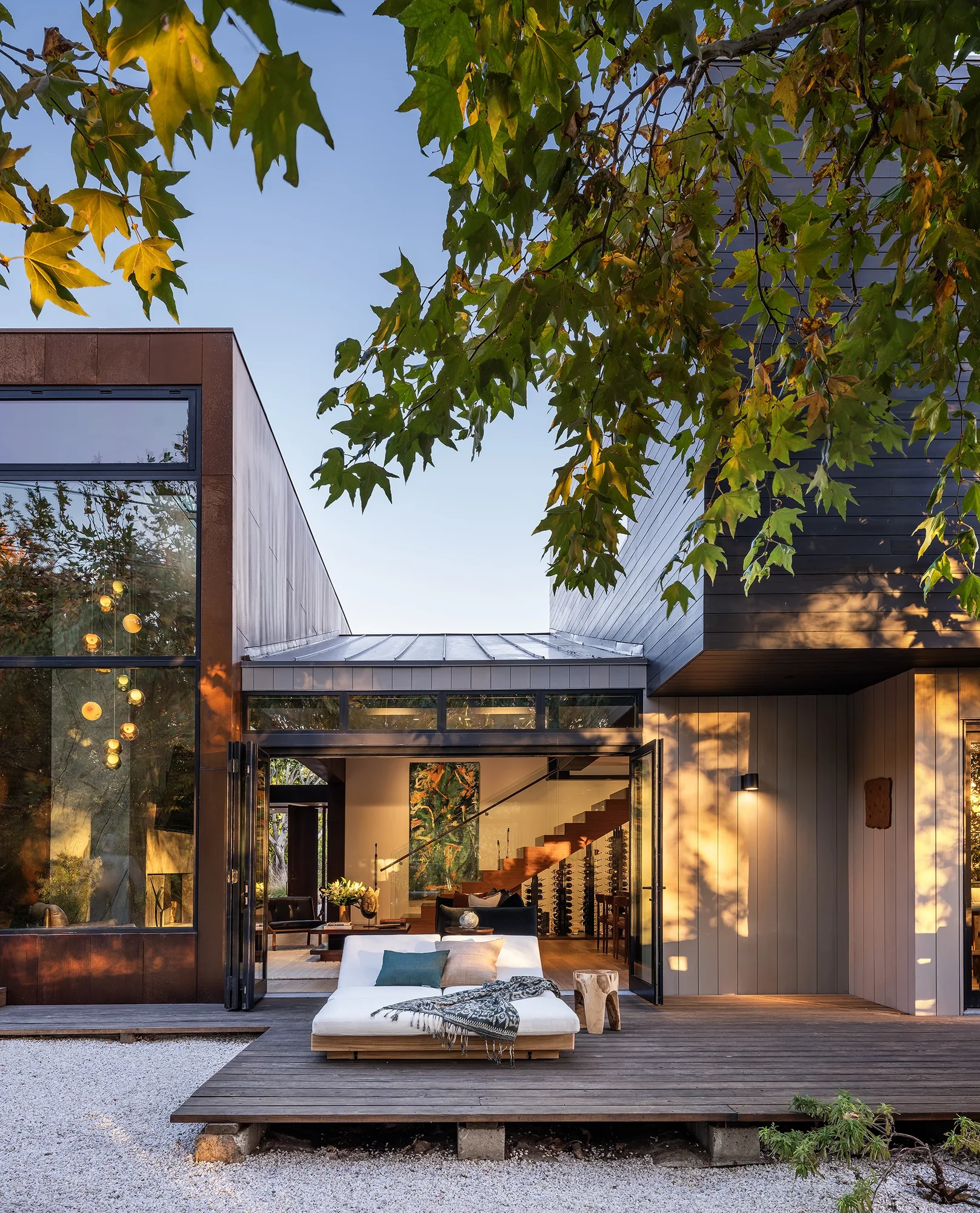 Modern home exterior with open sliding doors connecting wood deck to interior, framed by autumn tree canopy at dusk.