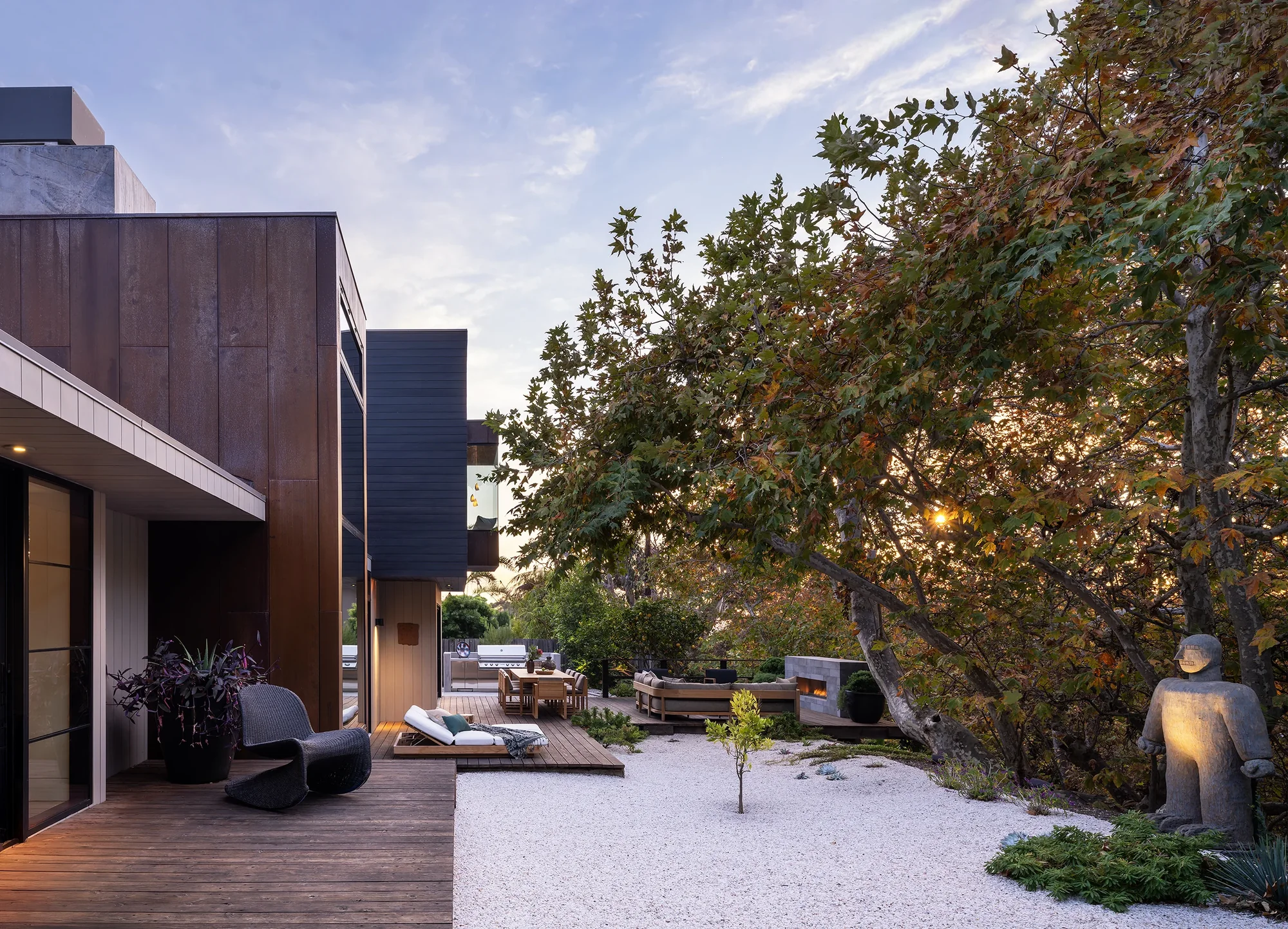 Modern outdoor living space with wood deck, gravel garden, and mature trees at dusk beneath open sky.