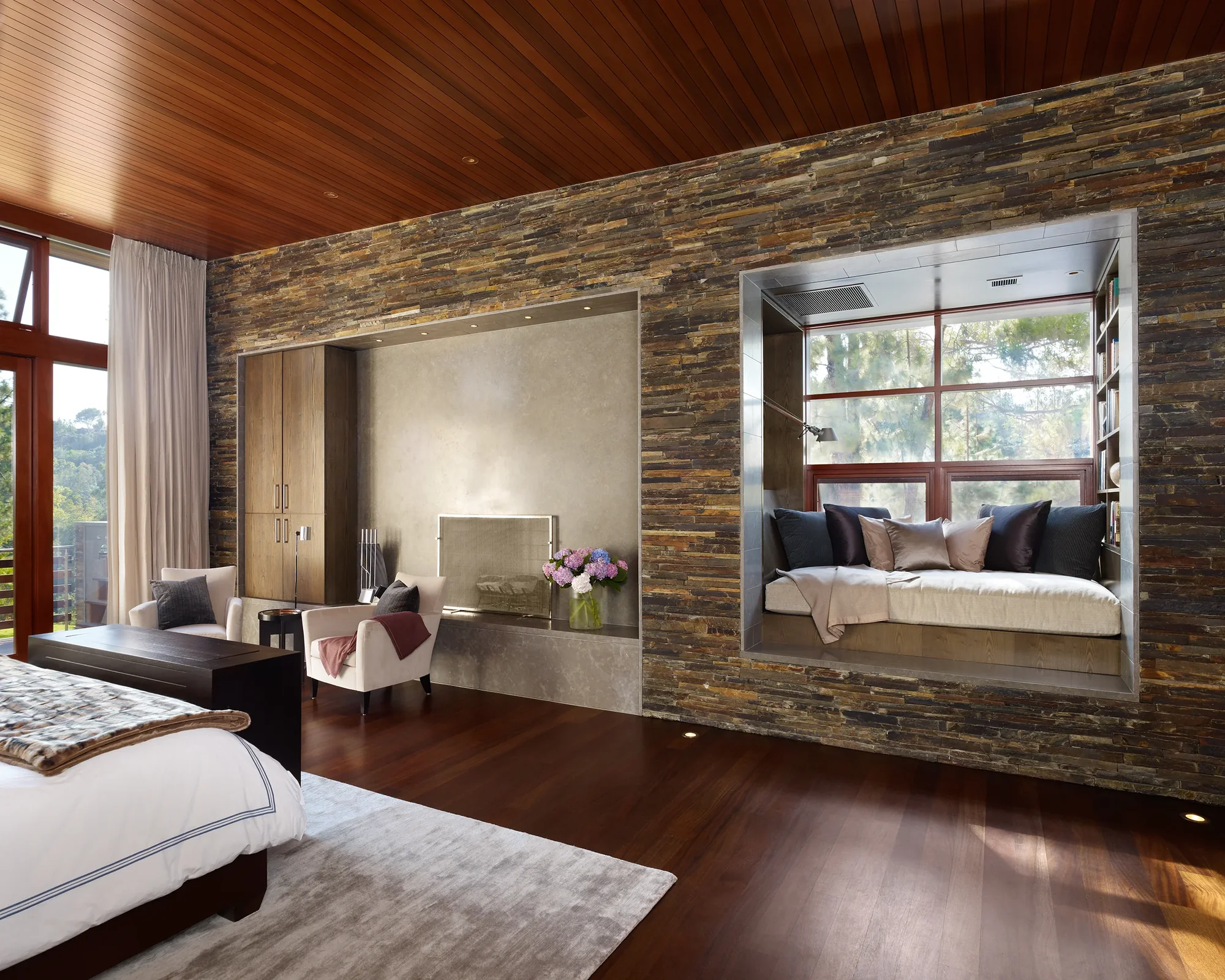Luxurious master bedroom with stacked stone wall, mahogany ceiling, built-in window seat, and floor-to-ceiling views of surrounding trees.