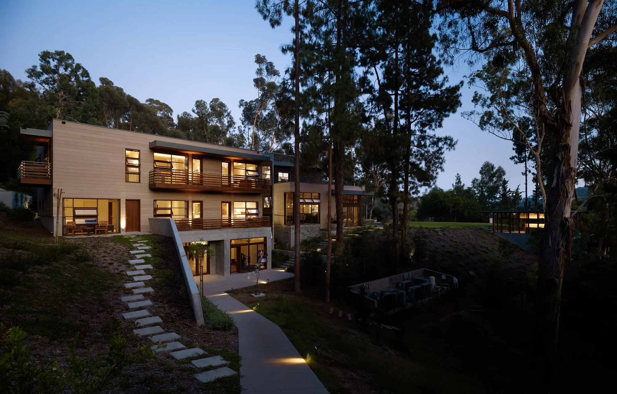 Illuminated modern home at dusk nestled among tall pines, with warm-lit windows, wood balconies, and stone pathways.