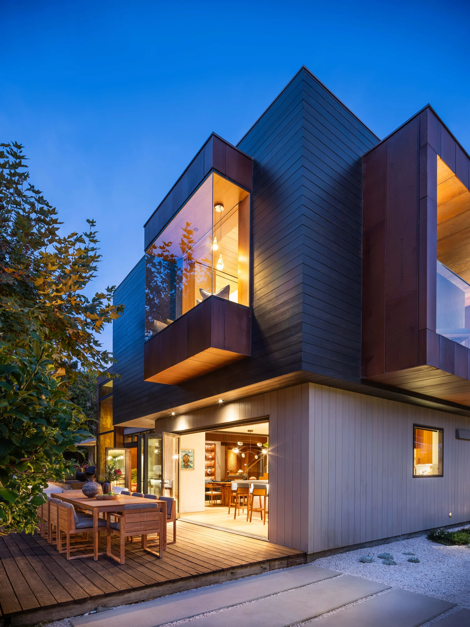 Modern two-story custom home at dusk with cedar siding, outdoor dining deck, and seamless indoor-outdoor connection