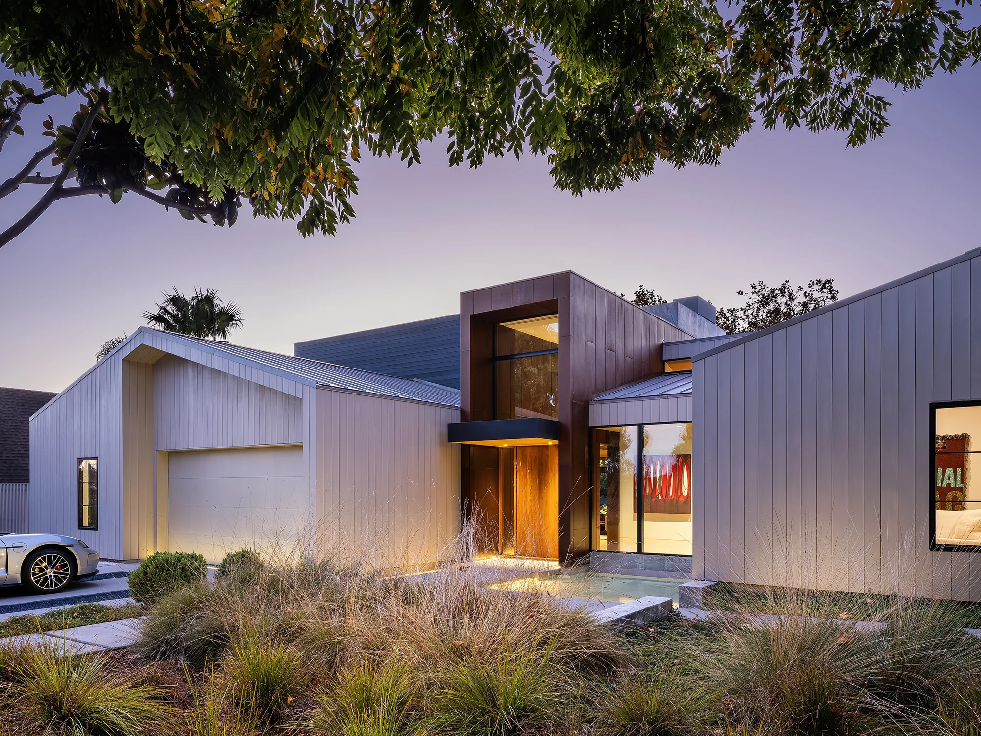 Modern single-story home with vertical cedar siding, illuminated entrance, and native grasses at dusk
